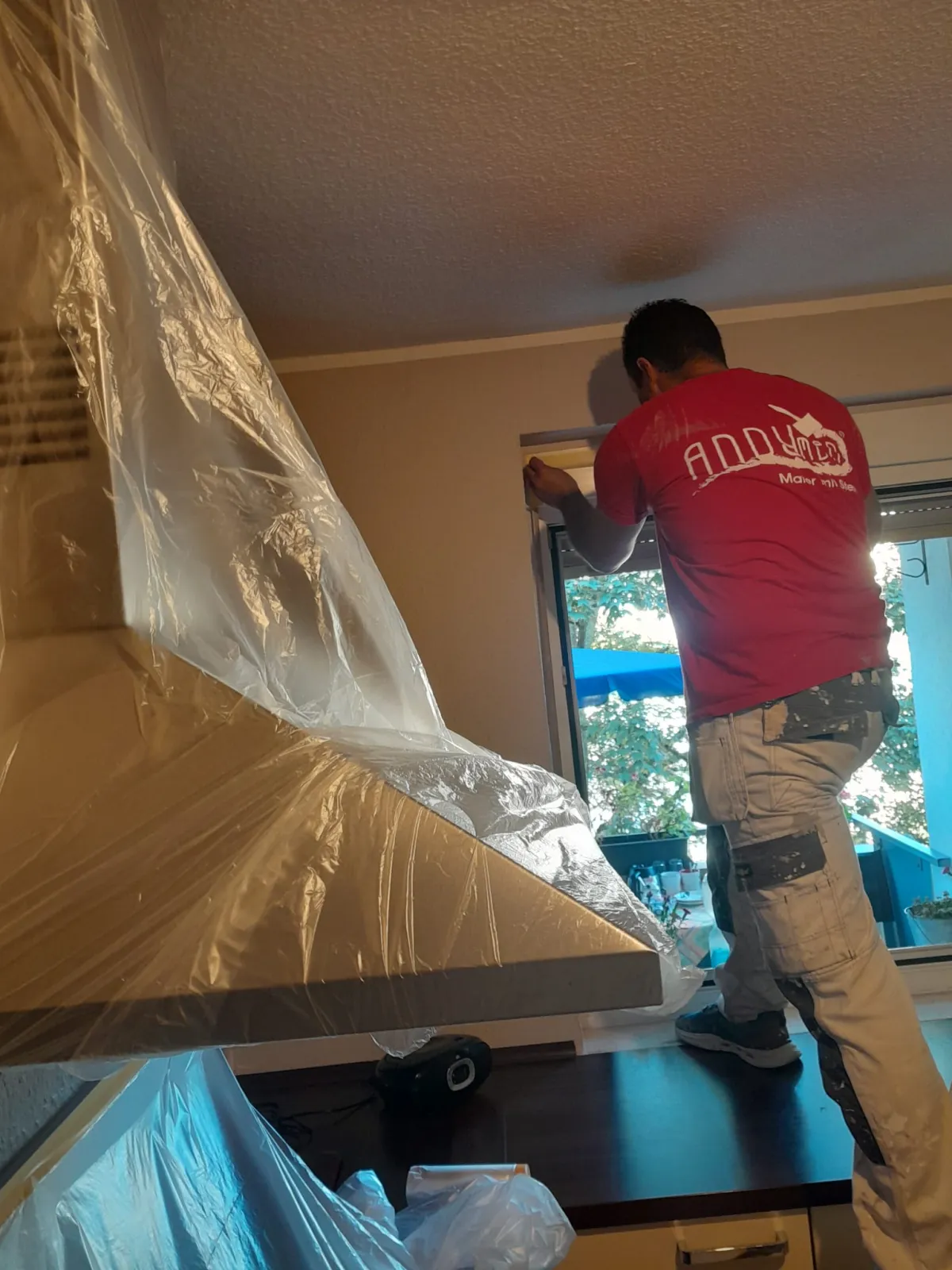 Maler klebt Fenster ab in K&uuml;che Andymen-Shirt von hinten sichtbar